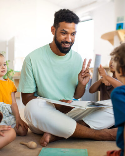 A male teacher holds up two fingers to group of children.